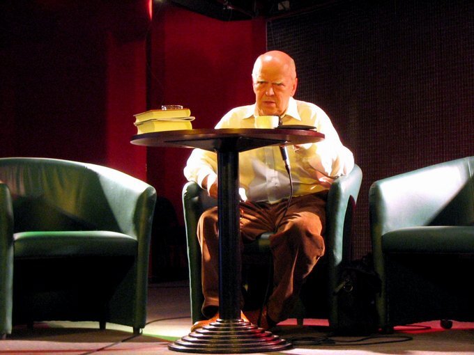 Zdjęcie przedstawia scenę, a na niej trzy stojące obok siebie fotele. Na środkowym fotelu siedzi starszy, łysy mężczyzna o owalnej twarzy. Ubrany jest w koszulę i spodnie. Przed mężczyzną stoi niewielki, okrągły stolik, na którym leżą książki i stoi filiżanka.