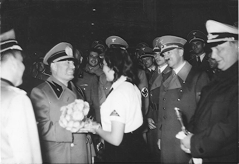 Zdjęcie przedstawia grupę niemieckich i hiszpańskich żołnierzy. Wśród nich są Adolf Hitler, Heinrich Himmler i Francisco Franco. Francisco Franco otrzymuje od młodej kobiety bukiet kwiatów.
