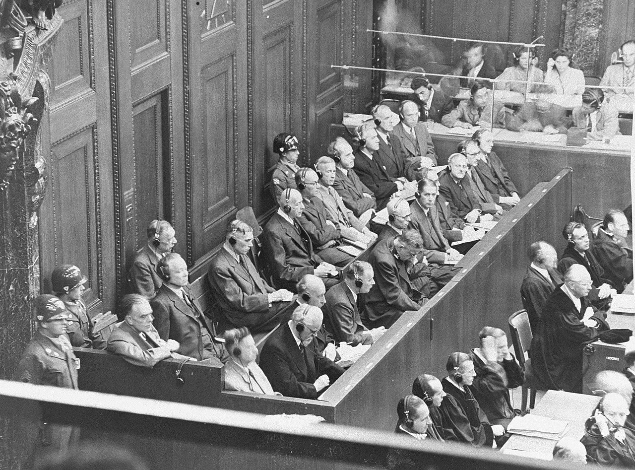 Zdjęcie przedstawia salę sądową. Przy kilku rzędach drewnianych ław siedzą mężczyźni i dwie kobiety. Większość ma założone słuchawki.