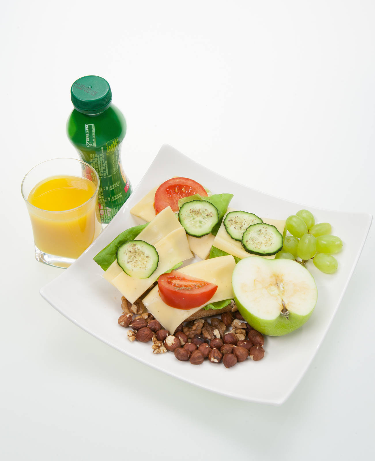 Fotografia przedstawia przygotowany posiłek śniadaniowy. Na talerzu znajdują się kanapki z pełnoziarnistego pieczywa, z serem i warzywami, a także owoce i orzechy. Obok talerza stoi plastikowa butelka z jogurtem, a także szklanka napełniona sokiem owocowym.
