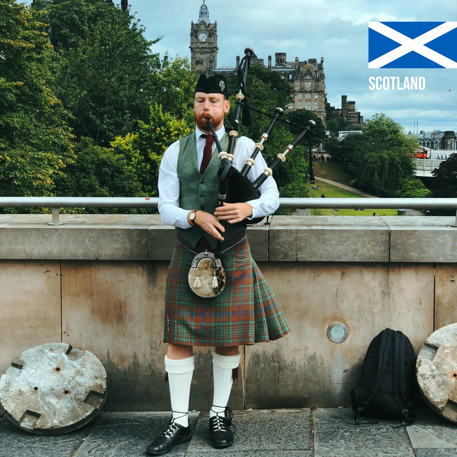 Zdjęcie przedstawia szkockiego kobziarza, od pasa w górę, ubranego w tradycyjny strój szkocki, który składa się z białej koszuli z długim rękawem, zielonej kamizelki, bordowego krawata. Na głowie mężczyzny widoczna jest czarna furażerka. Mężczyzna pod lewą pachą, w lewej ręce trzyma kobzę, zaś prawą ręką naciska na jej dolną część. W ustach ma jeden z ustników, w który dmie. Mężczyzna stoi na moście. W tle widoczne są zielone drzewa oraz kamienny budynek z wieżą zegarową. W prawym górnym rogu widoczna jest szkocka flaga.
