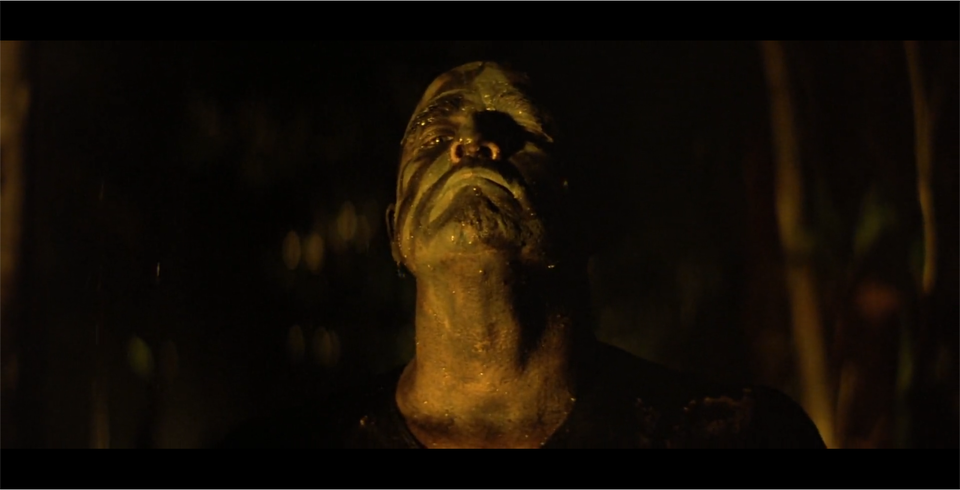 Kadr przedstawia łysego mężczyznę, który ma twarz pomalowaną na ciemnozielony kolor z ciemniejszymi pasami. Mężczyzna ma podniesioną głowę i ściśnięte usta. Patrzy przed siebie. Ubrany jest w ciemną koszulkę. W tle znajduje się las. Panuje noc.