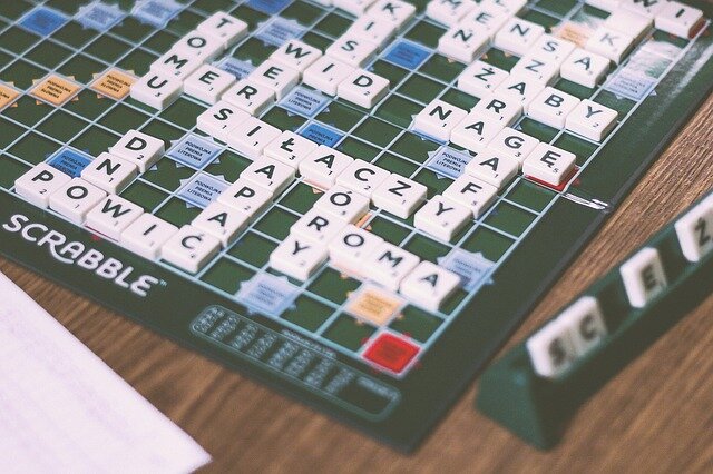 Fotografia przedstawia fragment planszy do gry scrabble. Gra leży na blacie i jest częściowo wypełniona wyrazami w języku polskim. Po prawej stoi plastikowa ramka z kilkoma kafelkami do gry.