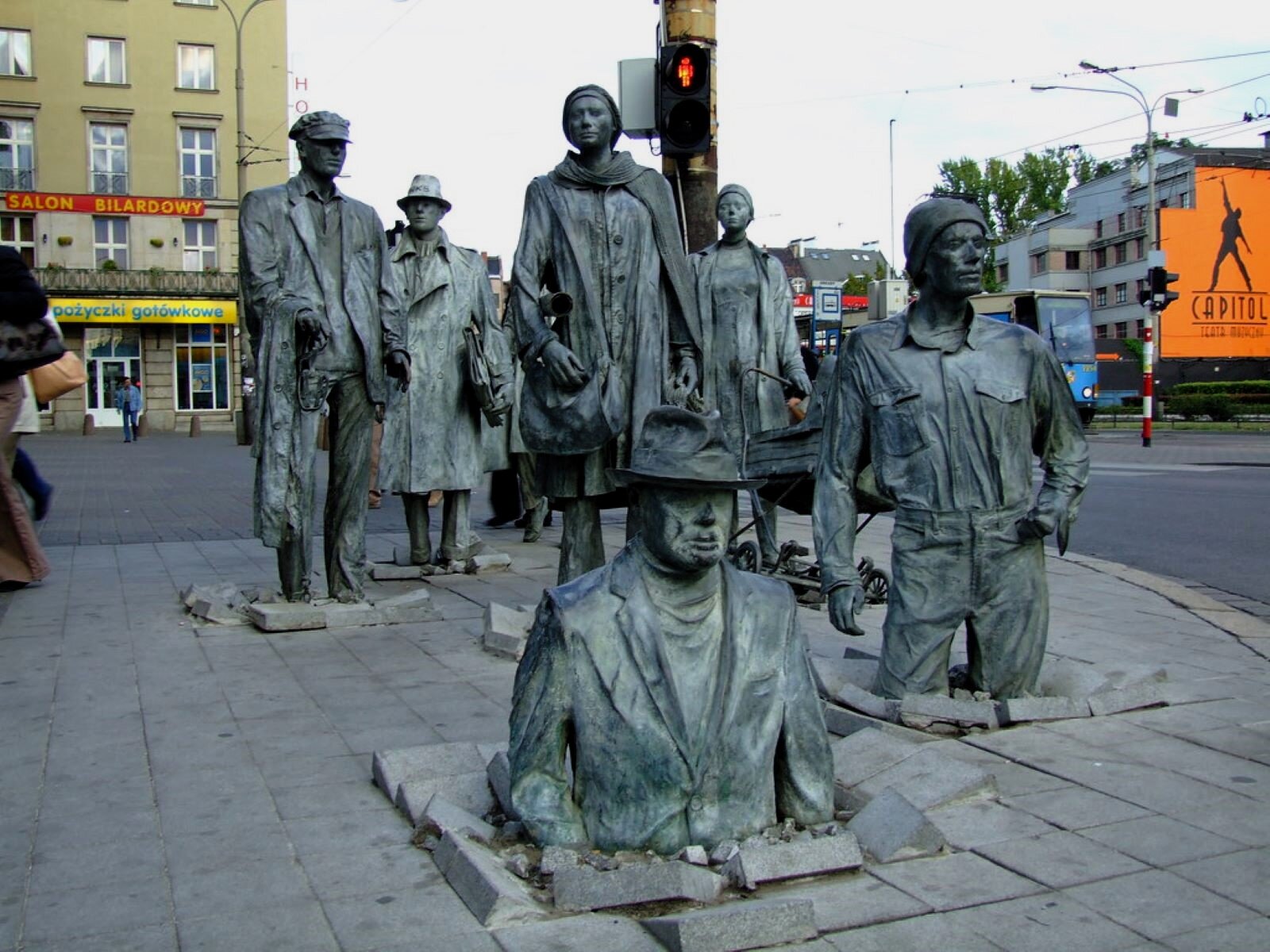 Zdjęcie przedstawia 14 odlanych w brązie figur  naturalnej wielkości, przedstawiających zwykłych ludzi, w codziennych ubraniach, z codziennym rekwizytami, wychodzącymi z chodnika w centrum Wrocławia. Instalację wykonał polski artysta Jerzy Kalina. 