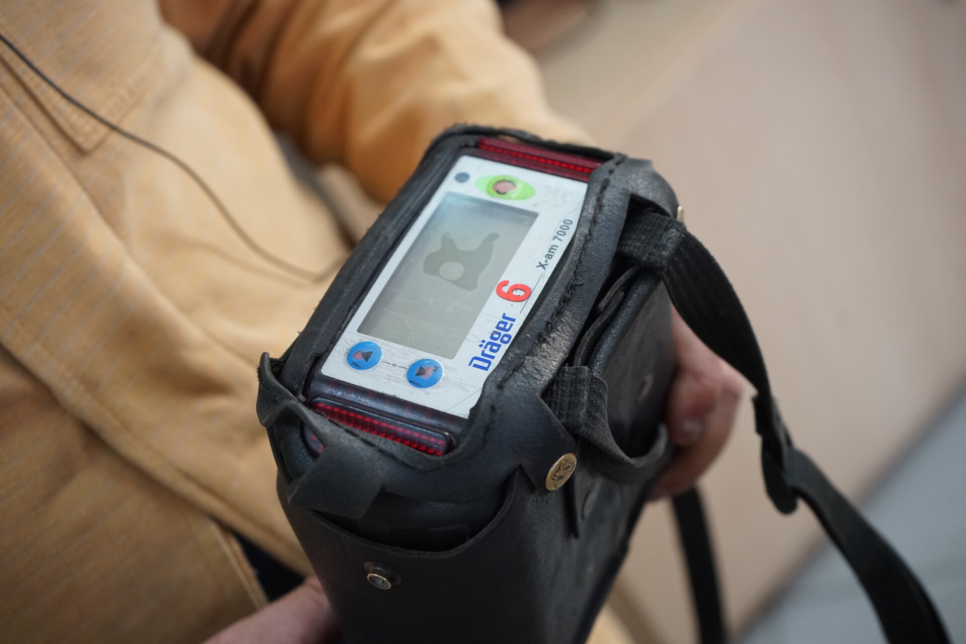 Zdjęcie przedstawia detektor do wykrywania do pięciu różnych gazów.  Detektor jest urządzeniem o kształcie prostopadłościanu. Górna podstawa prostopadłościanu składa się z czterech  przycisków oraz ekranu. Ekran zajmuje większą część powierzchni podstawy. Po lewej stronie widoczne są okrągłe przyciski z symbolami: grot strzałki w górę, grot strzałki w dół. Po prawej znajdują się niebieski okrągły przycisk oraz zielony przycisk. Na dole widnieje nazwa producenta oraz numer seryjny.Detektor znajduje się w pokrowcu. Detektor trzyma w dłoniach górnik.