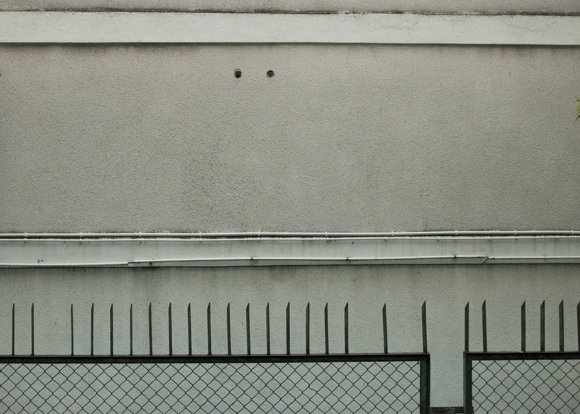 Ilustracja przedstawia zdjęcie jasnobeżowej elewacji budynku przedzielonej dwoma, biegnącymi poziomo, białymi gzymsami. U dołu kadru znajduje się metalowe ogrodzenie z siatką, zakończone ostrymi, pionowymi prętami. Statyczna kompozycja utrzymana jest w wąskiej, monochromatycznej, beżowej tonacji.