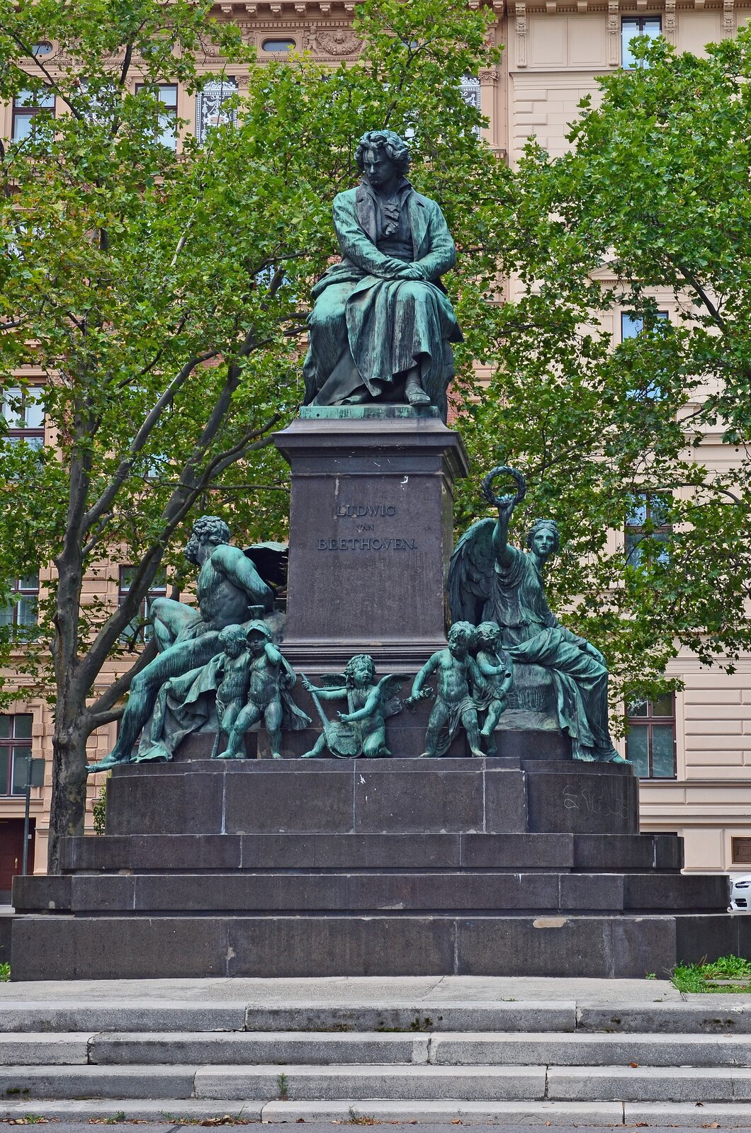 Ilustracja przedstawia pomnik z siedzącym Ludvigiem van Beethovenem w Wiedniu. Mężczyzna w średnim wieku, ubrany w szaty dostosowane do epoki. Włosy lekko kręcone, do ramion. Poniżej kompozytora znajdują się postaci ze skrzydłami.
