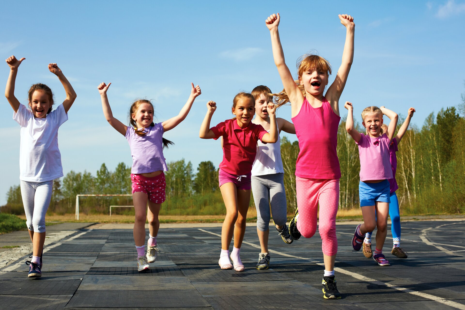 Zdjęcie przedstawia grupę dzieci biegnących po sportowej bieżni, z rękami uniesionymi do góry, w sportowych, kolorowych strojach i sportowych butach. Na pierwszym planie jest rudowłosa dziewczynka z kucykiem i grzywką. Za nią w drugiej linii, po lewej stronie biegną trzy dziewczynki, w trzeciej linii po prawej stronie chłopiec i dziewczynka. Za dziewczynką biegnie jeszcze jedno dziecko, które widoczne jest tylko częściowo. Na dalszym planie widać trawiaste boisko do piłki nożnej z dwoma metalowymi bramkami, a za nim las.