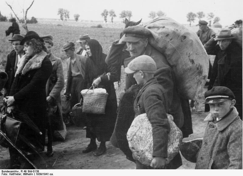 Zdjęcie przedstawia grupę ludzi idących drogą. Każda z osób dźwiga ze sobą bagaż. Kobieta w kapeluszu, ubrana w płaszcz z kołnierzem z futra, niesie torebkę i parasol. Kobieta za nią w chuście na głowie niesie siatkę. Dwaj mężczyźni niosą worki na plecach. Chłopiec trzyma pod pachą zawiniątko. Za placami jednego z mężczyzn wychyla się końska głowa. Koń ciągnie wóz. W tle pola i pojedyncze drzewa. 