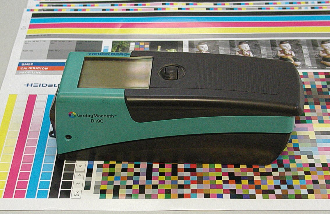 Zdjęcie przedstawia densytometr położony na wydrukach z profilami barwnymi, próbkami kolorystycznymi, skalą szarą. Widoczna jest obudowa przyrządu o prostopadłościennym kształcie, w kolorze czarnym i zielonym. Widoczny jest dłuższy bok i góra. W górnej części znajduje się wyświetlacz oraz pokrętło do jego obsługi.