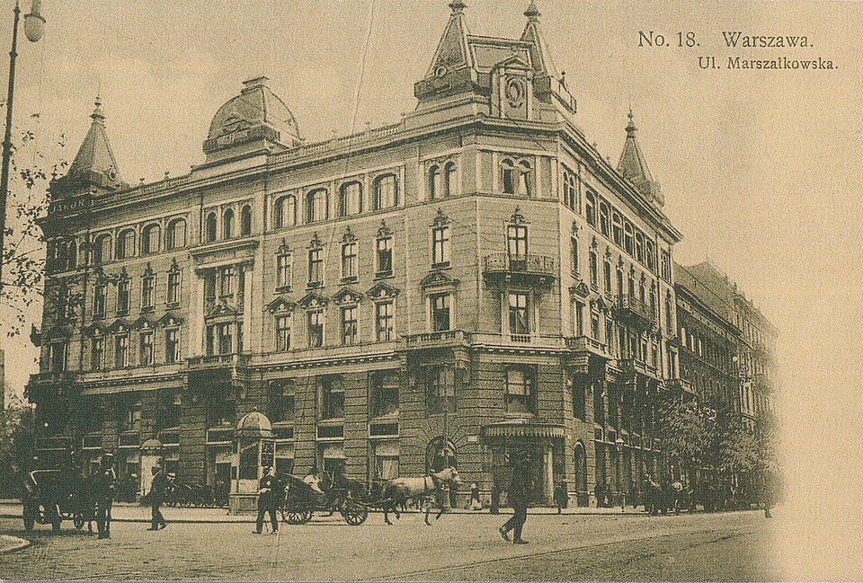Archiwalna pocztówka przedstawiająca Domu Mody Bogusława Hersego w przedwojennej Warszawie. Na pierwszym planie widać szerokie skrzyżowanie, po którym poruszają się konne powozy i dorożki. Ulicą spacerują nieliczni przechodnie ubrani w stroje z przełomu XIX i XX wieku. W prawym górnym rogu widnieje podpis identyfikujący lokalizację oraz numer serii wydawniczej.

