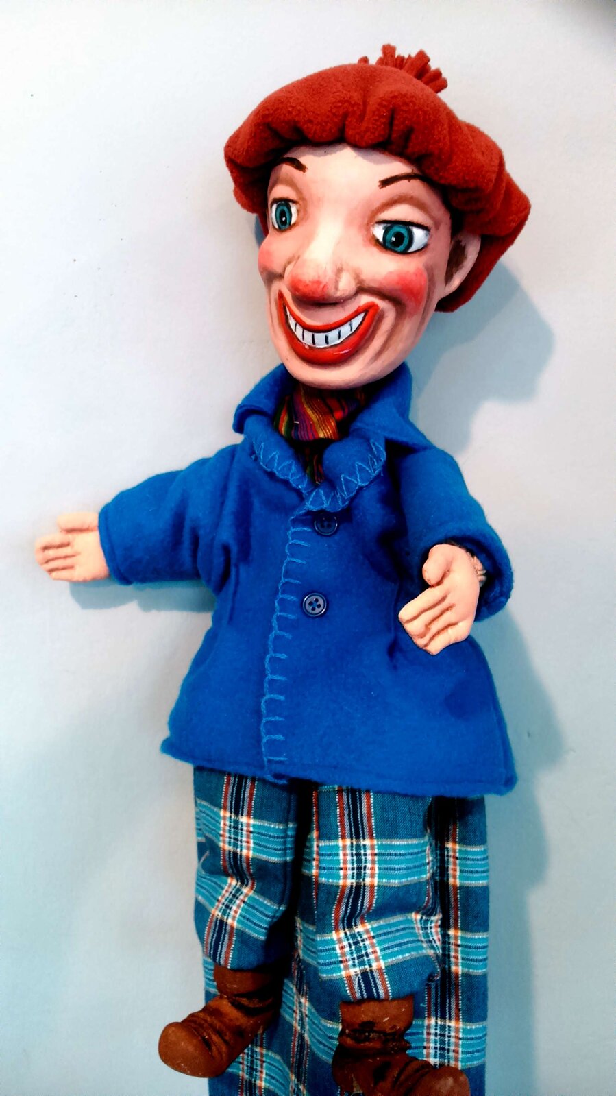 Ilustracja przedstawia uśmiechniętą pacynkę. Lalka to postać chłopca ubranego w niebieski płaszczyk oraz kraciaste spodnie. Na nogach ma brązowe trzewiki.