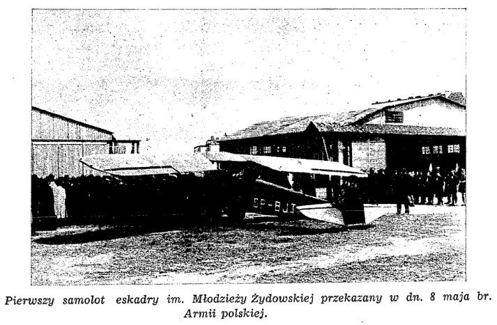 Zdjęcie przedstawia samolot. Za samolotem widoczne są hangary - budynki gospodarcze. Po prawej stronie stoi grupa ludzi.