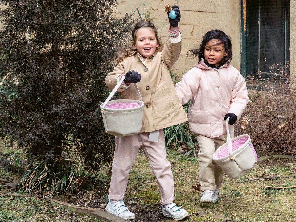 Zdjęcie przedstawia dwie dziewczynki w wieku wczesnoszkolnym, które idą po wysuszonej trawie i trzymają w dłoniach koszyczki.  Jedna z dziewczynek trzyma w wysoko uniesionej dłoni jajko. 
