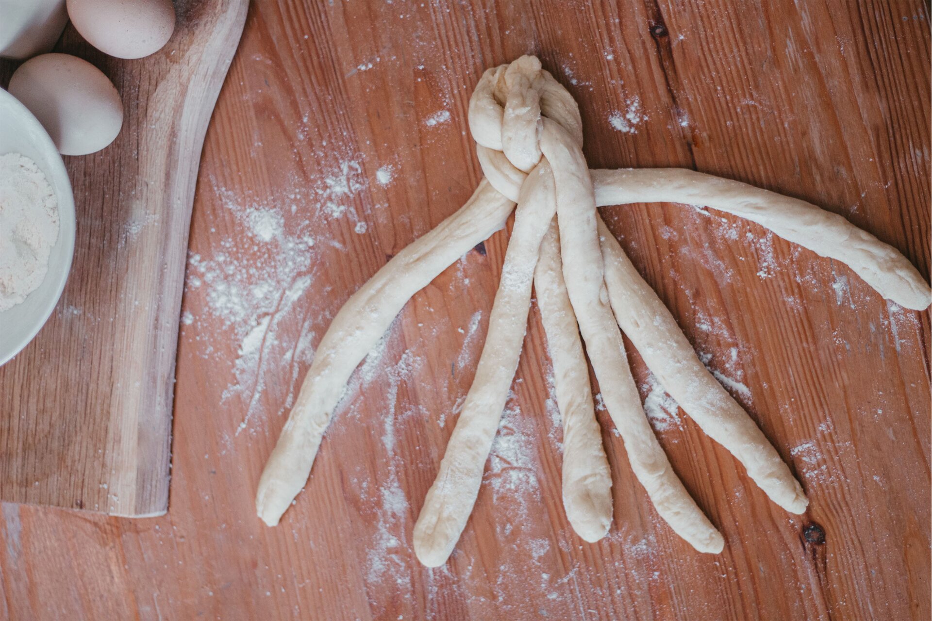 Zdjęcie przedstawia krok czwarty i piąty formowania chały kijowskiej. Ujęcie wykonane jest od góry pod skosem. Widoczny jest ciemnobrązowy drewniany blat obsypany białą mąką. Na nim w środkowej części grafiki znajdują się wałki ciasta, które są przez siebie wzajemnie przeplecione. W lewej części zdjęcia dostrzec można drewnianą jasnobrązową deskę ułożoną pionowo. Na niej znajduje się biała miseczka wypełniona do połowy mąką. Nad nią dostrzec można dwa jajka.