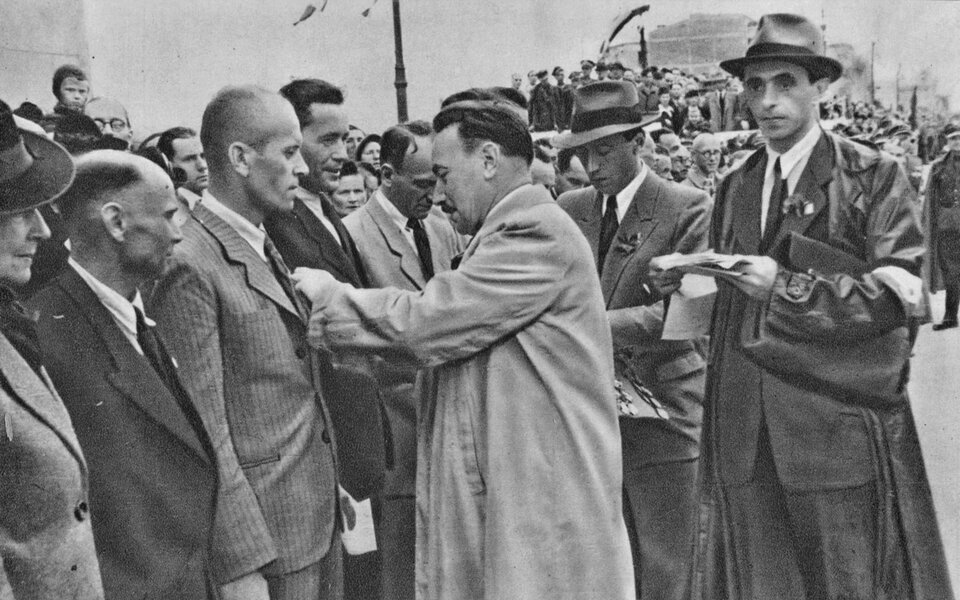 Ilustracja przedstawia kilkanaście osób stojących w szeregu. Przed nimi mężczyzna w jasnym płaszczu nachyla się, by zawiesić na połach marynarki odznaczenie jednej z osób. Obok niego, dwaj mężczyźni w kapeluszach, którzy trzymają w rękach odznaczenia dla kolejnych osób. W tle tłumu widoczne są polskie flagi. 