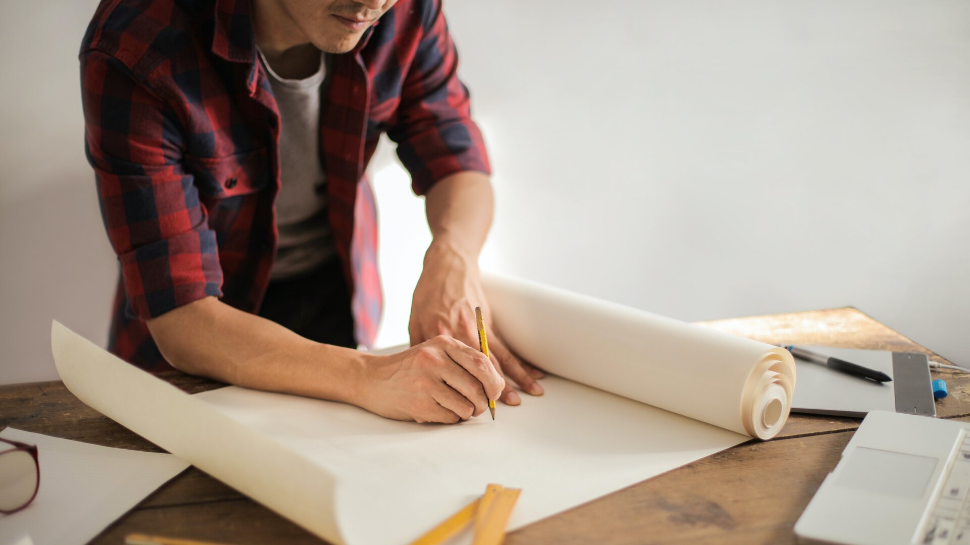 Ilustracja przedstawia mężczyznę stojącego nad stołem i rysującego ołówkiem na rozwiniętym zwoju papieru. Mężczyzna ma na sobie rozpiętą koszulę w czerwono‑granatową kratę i białą koszulkę. Na stole oprócz zwoju papieru leżą także: linijka, długopis, tablet, karta i okulary przeciwsłoneczne.