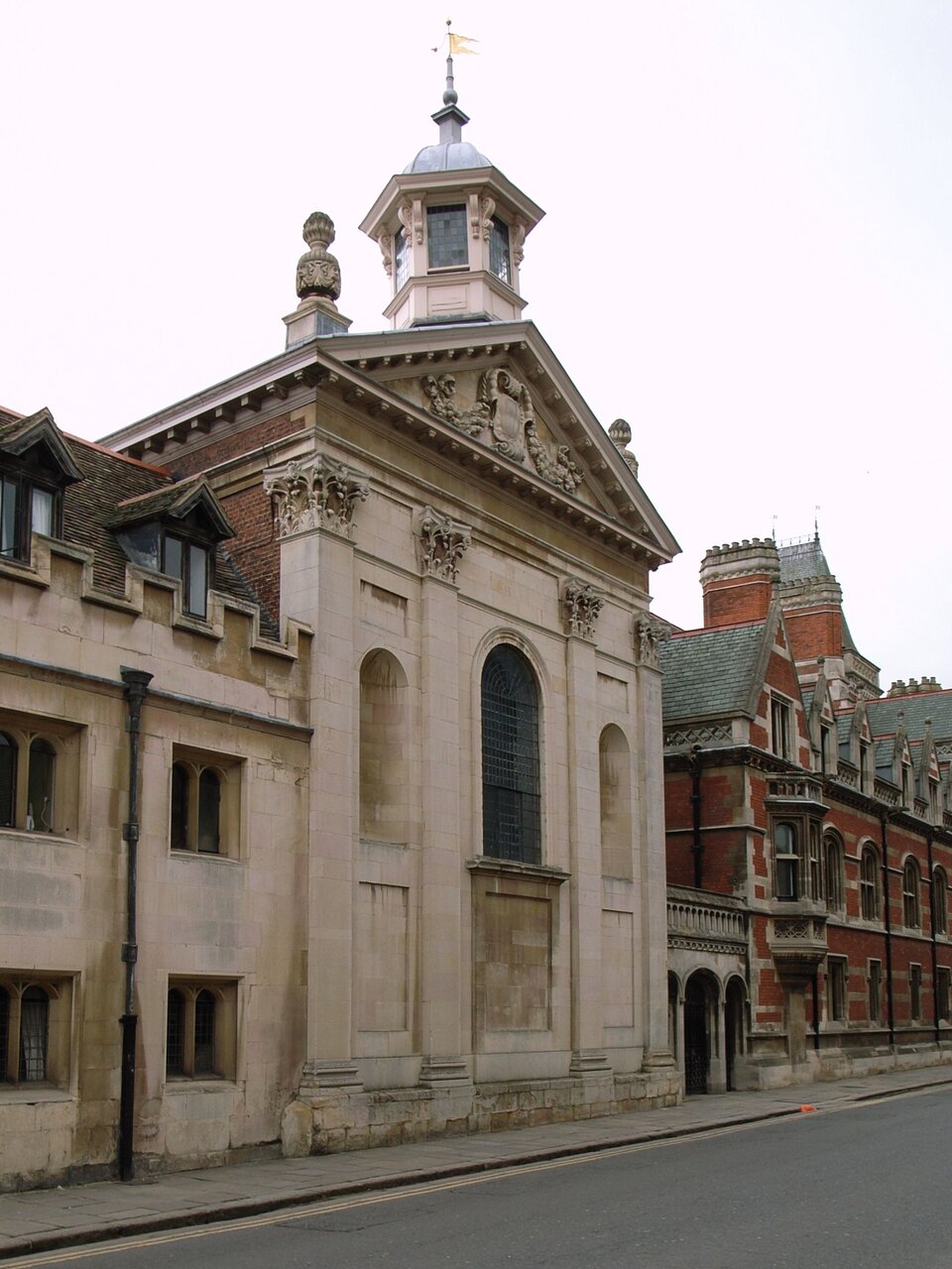 Ilustracja przedstawia zdjęcie kaplicy w Pembroke College. W kadrze znajduje się zwarta zabudowa wzdłuż ulicy. Wśród budynków wyróżnia się kaplica: jest najwyższa, jej fasada jest w jasnym kolorze. Fasad ma układ wertykalny, który podkreślają cztery pilastry od podstawy do fryzu budynku. Nad gzymsem znajduje się tympanon wypełniony płaskorzeźbą. Między pilastrami są trzy wnęki zwieńczone łagodnym łukiem, z czego środkowa, największa - stanowi okno, pozostałe rozmieszczone symetrycznie, po jego obu stronach – są ślepymi oknami.