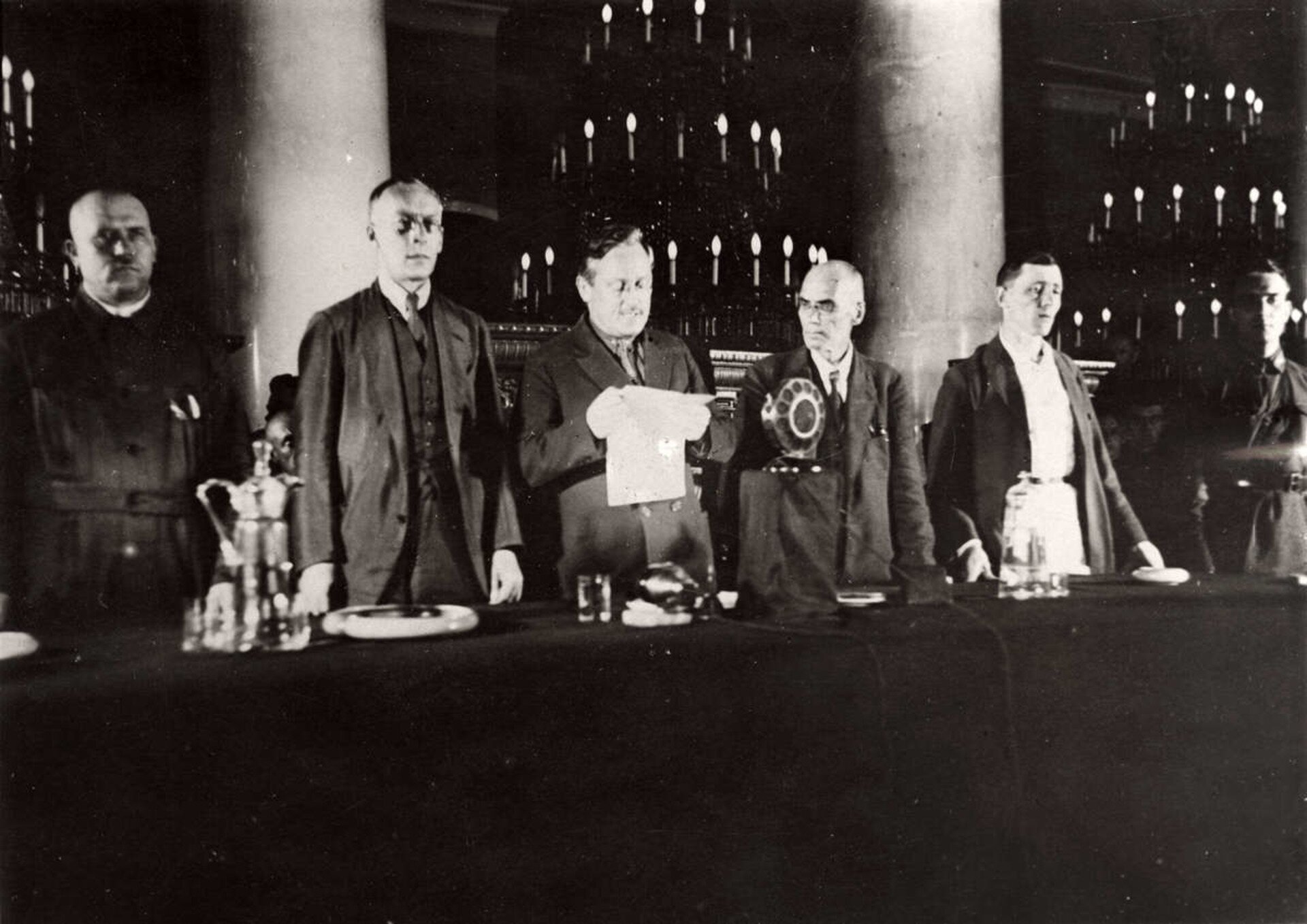 Fotografia czarno-biała przedstawiająca Andreygo Vyshinsky, który odczytuje akt oskarżenia. Pozostali członkowie stoją obok niego i słuchają odczytu patrząc w tłum.