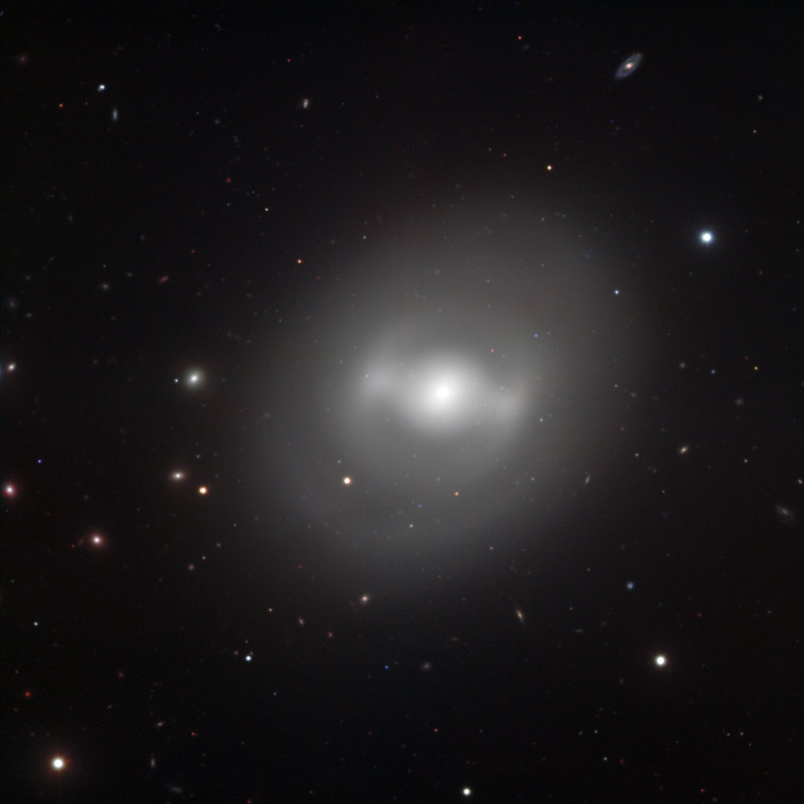 Grafika przedstawia galaktykę soczewkowatą z poprzeczką NGC 936. Galaktyka to jasny, położony centralnie punkt z jasną poświatą dookoła oraz małymi, jasnymi punktami na czarnym tle.   