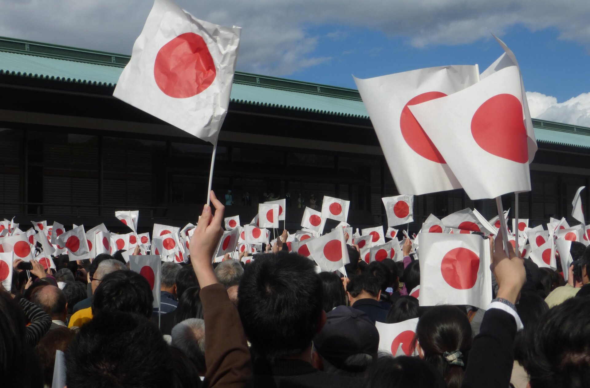 Na zdjęciu znajduje się tłum ludzi, wszyscy trzymają w rękach flagę Japonii. Flaga ma białe tło a na jej środku znajduje się czerwone koło. 