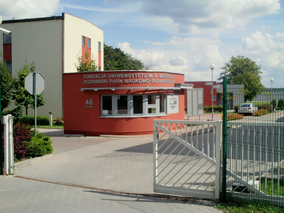 Na zdjęciu widoczny jest wjazd na teren Poznańskiego Parku Naukowo‑Technicznego. Biała rozsunięta brama znajduje się na pierwszym planie. Nawierzchnia pokryta jest szarą kostką.  Na drugim planie jest kompleks nowoczesnych budynków. W centralnej części ciemnopomarańczowa niska rotunda z szarymi oknami i szarym napisem drukowanymi literami Fundacja Uniwersytetu im. A. Mickiewicza Poznański Park Naukowo‑Techniczny, a poniżej po prawej stronie numer 46 i niewidoczna nazwa ulicy. Po prawej stronie rotundy fragment kremowego dwupiętrowego budynku, a po prawej niskie budynku i parking z widocznym jednym samochodem. Przy drodze dwie latarnie i niewielkie ilości zieleni. W tle niebiesko‑szare lekko zachmurzone niebo.