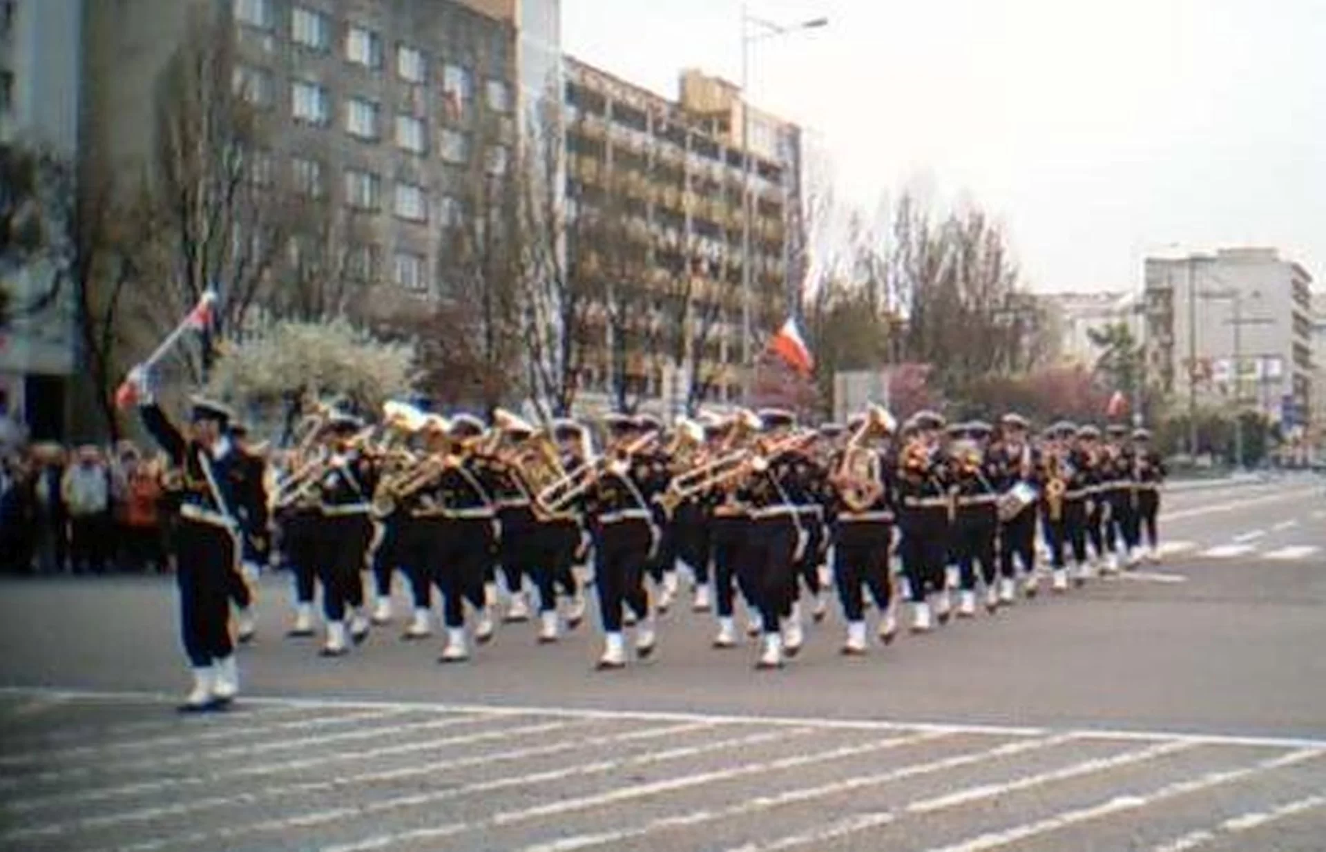 Ilustracja przedstawia orkiestrę dętą typu fanfara. Muzycy ubrani są w czarne mundury, białe czapki, pasek i buty. Maszerują ulicą. W tle widać bloki i tłum ludzi.