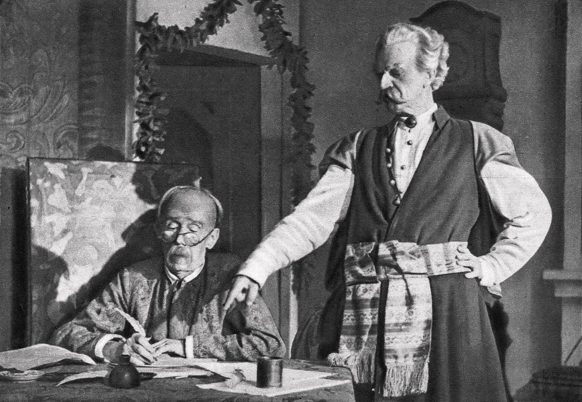 Czarno‑biała fotografia przedstawia starszych mężczyzn, znajdujących się w paradnej komnacie szlacheckiego zamku. Jeden z nich siedzi przy stole i pisze list gęsim piórem. Ma skupioną na czynności owalną twarz, jest łysy, ma cienkie brwi, prosty nos i sumiaste wąsy. Zza okrągłych okularów spogląda na zapisaną kartką papieru. W prawej dłoni trzyma pióro. Na stole leżą kartki papieru i stoi kałamarz z atramentem. Mężczyzna nosi żupan. Obok stoi drugi mężczyzna ubrany w żupan i kontusz przewiązany pasem słuckim w poziome paski, ozdobiony frędzlami. Szlachcic stoi wyprostowany i prawą ręką z wyciągniętym palcem wskazującym pokazuje na zapisaną kartkę. Ma on owalną twarz, sumiaste wąsy, gęste, lekko falujące, siwe włosy z zakolami. Wyraz jego twarzy jest stanowczy, brwi zmarszczone, oczy głęboko osadzone, a nos haczykowaty. Lewą rękę opiera o biodro.