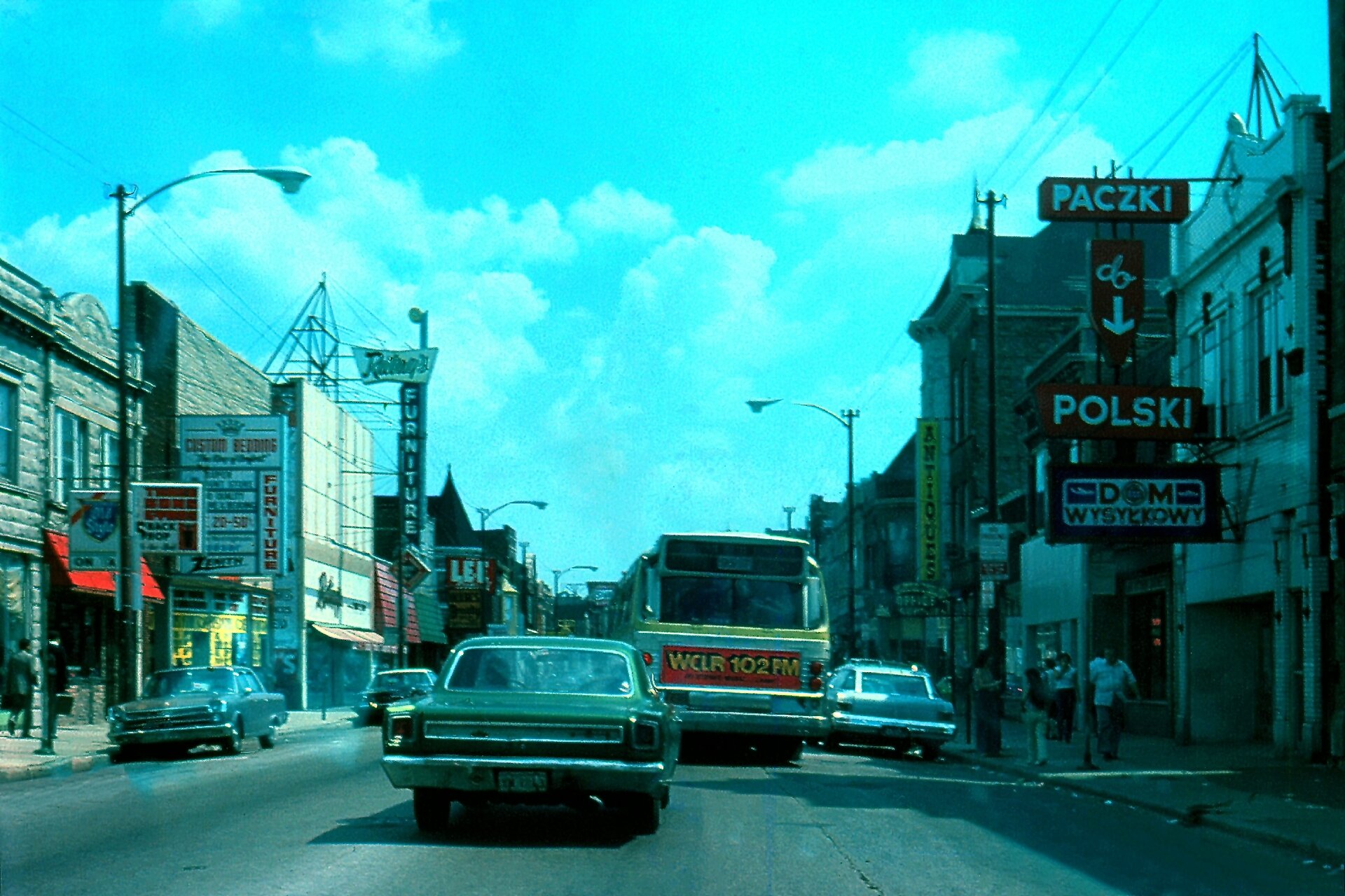 Na zdjęciu ulica w mieście. Liczne szyldy i reklamy z napisami w języku polskim.
