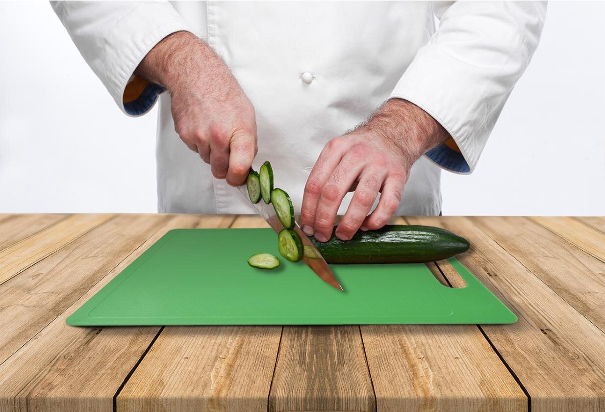 Zdjęcie przedstawia ogórek krojony techniką rouelle. Na drewnianym blacie widoczna jest zielona deska, na której znajduje się częściowo pokrojony w plastry ogórek. W tle postać ubrana w strój kucharza, który prawą ręką trzyma trzonek noża, którego ostrze dotyka ogórka i na którym znajduje się kilka pokrojonych plastrów. Lewa ręka postaci przytrzymuje lekko zgiętymi palcami część niepokrojoną ogórka.