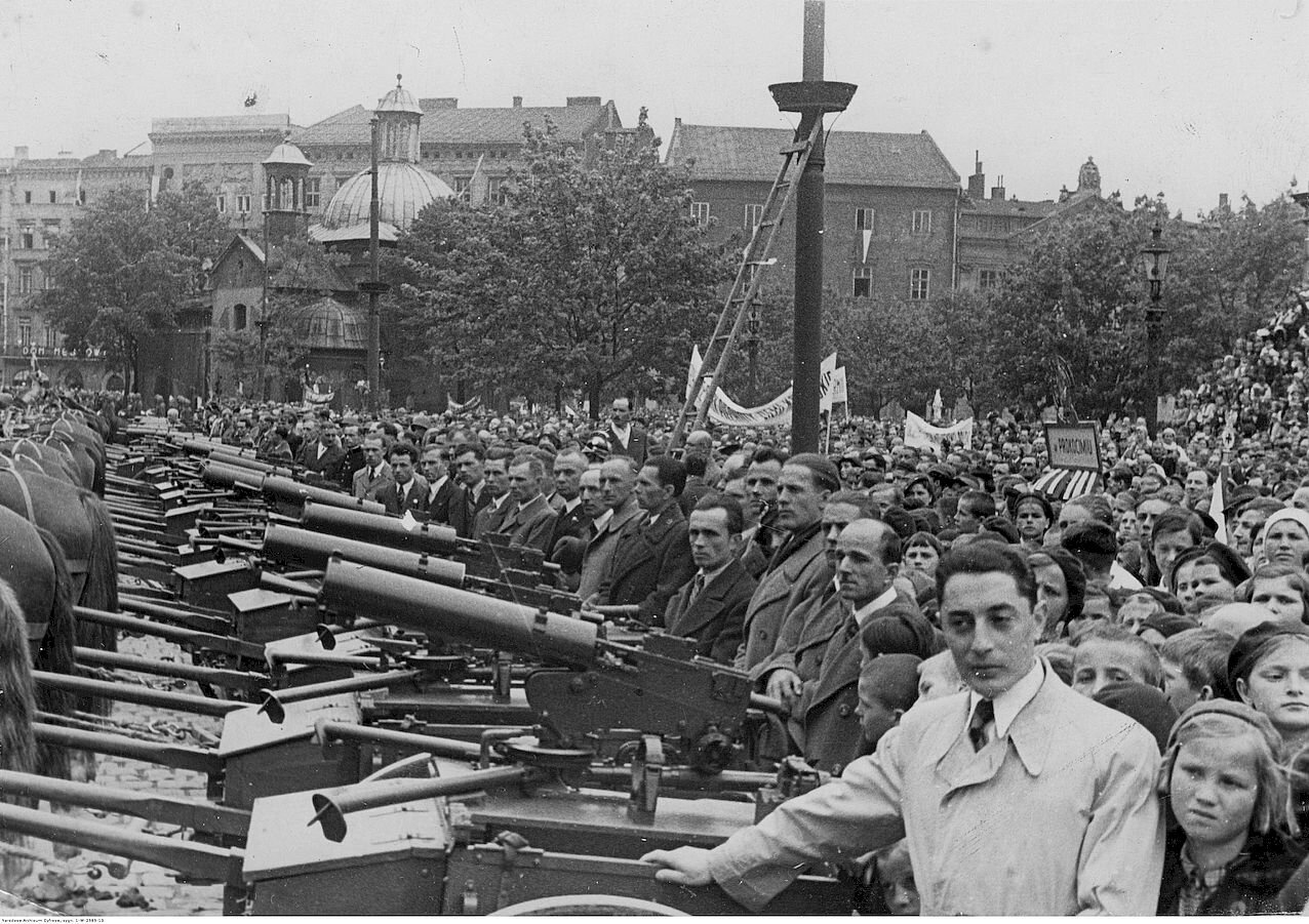 Zdjęcie przedstawia rząd armat ustawiony na placu miejskim. Po prawej stronie jest tłum ludzi.