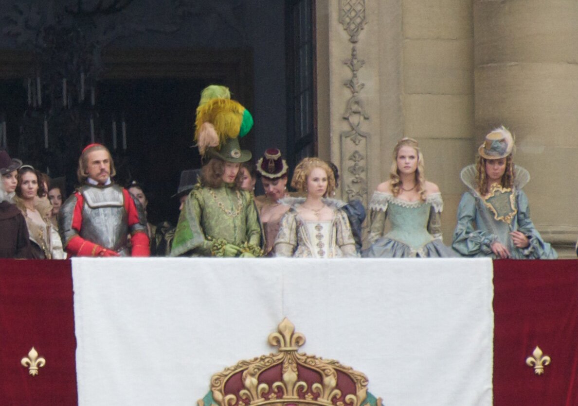 Kolorowa fotografia z adaptacji filmowej "Trzech muszkieterów" przedstawia króla Ludwika w otoczeniu dworu. Zebrani stoją na balkonie pałacu i na coś patrzą. Od lewej stoją: kardynał  Richelieu   w zbroi , król Ludwik XIII w zielonkawym stroju, na głowie ma kapelusz w wielkimi żółto‑zielonymi piórami, obok królowa Anna w srebrnej sukni, następnie jej dwórka Konstancja w zielonkawej sukni i dama dworu, w kapeluszu z piórem. Wszyscy opierają dłonie na balustradzie, na której zawieszono godło Ludwika XIII. Po obu stronach godła ciemnoczerwone pasy, na nich złote lilie, pośrodku biały pas, na nim złota korona.