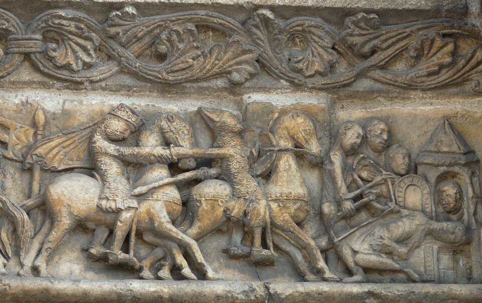 Zorientowana poziomo fotografia barwna przedstawiająca płaskorzeźbę, fragment fryzu z arkad fasady katedry Saint‑Pierre w Angoulême, przedstawiający sceny z Pieśni o Rollandzie. Kamień, w którym wykuto fryz, nosi znaki czasu i miejscami jest powykruszany. U góry kadru, powyżej dwóch trzecich jego wysokości, przebiega roślinny ornament. Poniżej wąski pas zaprawy wapiennej, a pod nią płaskorzeźba przedstawiająca w uproszczony sposób charakterystyczne dla sztuki romańskiej figuratywne przedstawienie. Poczynając od lewej na galopującym ku prawej stronie koniu, stojący w strzemionach jeździec z pochyloną do przodu głową w koronie, ubrany w łuskową zbroję sięgającą kolan, nagolenniki i ostrogi na piętach. Z korony na plecy spływa rozwiany krótki płaszcz. W ręce prawej wyciągniętej ku przodowi  trzyma poziomo ustawiony miecz. Sięga on ostrzem piersi podobnie odzianego drugiego poprzedzającego go jeźdźca z biskupią tiarą na odchylonej do tyłu głowie. Jeździec ten w odwiedzionej do tyłu prawej ręce dzierży włócznię, godząc udo podążającego za nim napastnika. Jednocześnie nad kark swojego konia unosi okrągłą tarczę, a podtrzymującą ją dłonią jednocześnie pociąga wodze, by nim kierować. Z prawej strony przed rumakiem trzy postaci pochylają się nad omdlałym ciałem mężczyzny. Ten na wznak z podkulonymi nogami spoczywa oparty plecami na siedzeniu tronu. Prawą dłonią przytrzymuje cięciwę i fragment złamanego łuku, którego drugą część trzyma w opuszczonej dłoni pierwsza postać od lewej ze wspomnianych trzech. W tle za głową leżącego w oknie zwieńczonej stożkowym dachem baszty, głowa jeszcze jednej osoby.