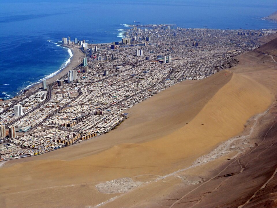 Zdjęcie przedstawia widok z lotu ptaka na znacznej wielkości nadmorskie miasto. Na górze i w lewym górnym rogu znajduje się morze, następnie mniej więcej od lewego dolnego rogu do prawego górnego rogu rozpościera się miasto. Pozostałą część zdjęcia wypełnia bardzo duża wydma paraboliczna. Jej piasek ma ciemnopomarańczowy kolor. Stok dowietrzny jest dużo łagodniejszy niż zawietrzny. Wydma jest wielkości porównywalnej z leżącym u jej podnóża miastem.
