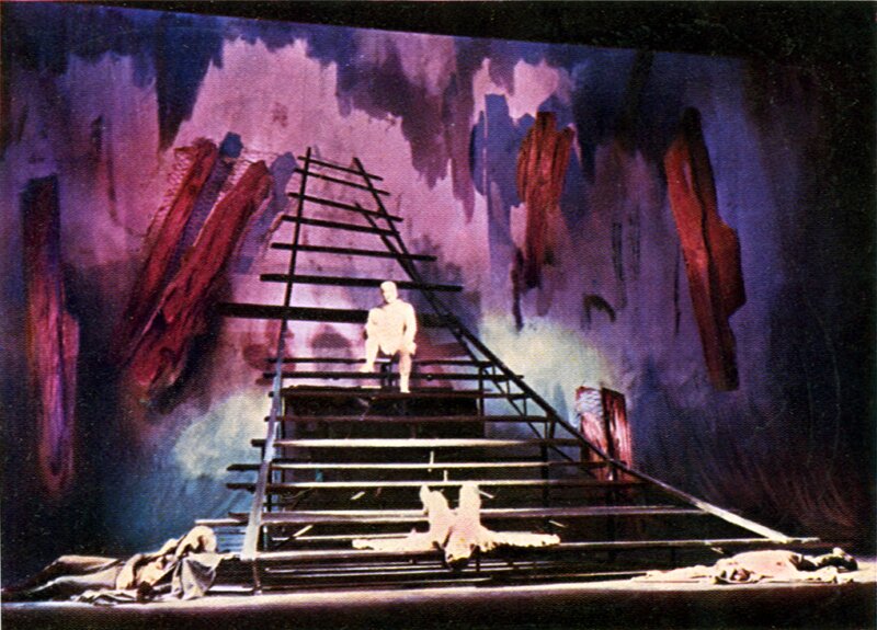 Fotografia przedstawia teatralną scenę. Pośrodku stoi drewniana konstrukcja składająca się w wielu zwężających się schodów. Jest ona zakrzywiona u góry. Na schodach siedzą i leżą postaci ubrane w proste szaty. W tle ukazano planszę ozdobioną czerwonymi plamami. 