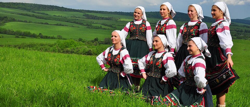 Ilustracja przedstawia Łemkowski Zespół Pieśni i Tańca „Kyczera”. Sceneria zdjęcia odgrywa się na łące na zielonej trawie. Kobiety ubrane w koszule z lnianego płótna. Do spódnicy noszono zapaskę, zdobioną kolorowym haftem. Na głowach mają zawiązane białe chusty. 