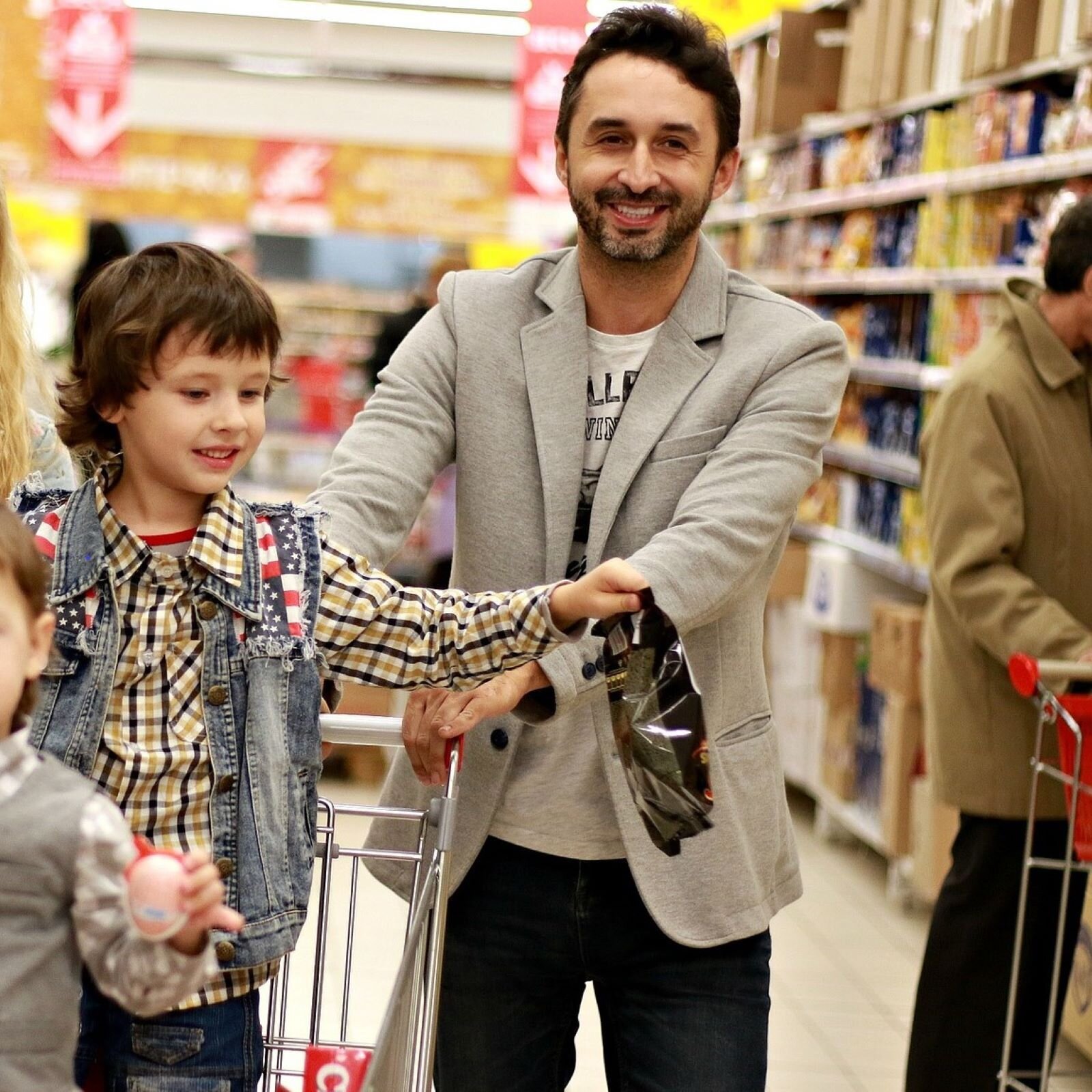 Zdjęcie przedstawia uśmiechniętego mężczyznę prowadzącego wózek w alejce sklepowej. W wózku stoi uśmiechnięty chłopiec w wieku około 4 lat.