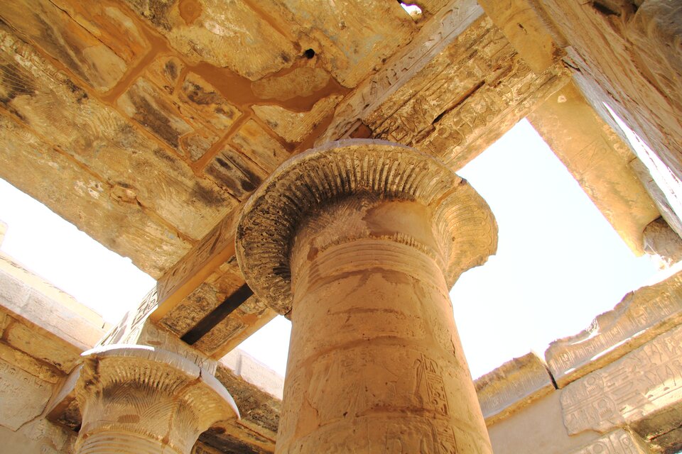 Fotografia ukazuje fragment wnętrza świątyni Chonsu w Karnaku- widok na masywne kolumny i kamienny strop. W centrum kadru dominuje kolumna o kapitelu w formie rozwiniętego kwiatu papirusu. Trzon kolumny zdobią płaskorzeźby oraz hieroglify. Kapitele są bogato rzeźbione, a ich powierzchnia nosi ślady dawnych kolorów. Ponad kolumnami rozciąga się architraw i belkowanie, również pokryte reliefami i inskrypcjami. 