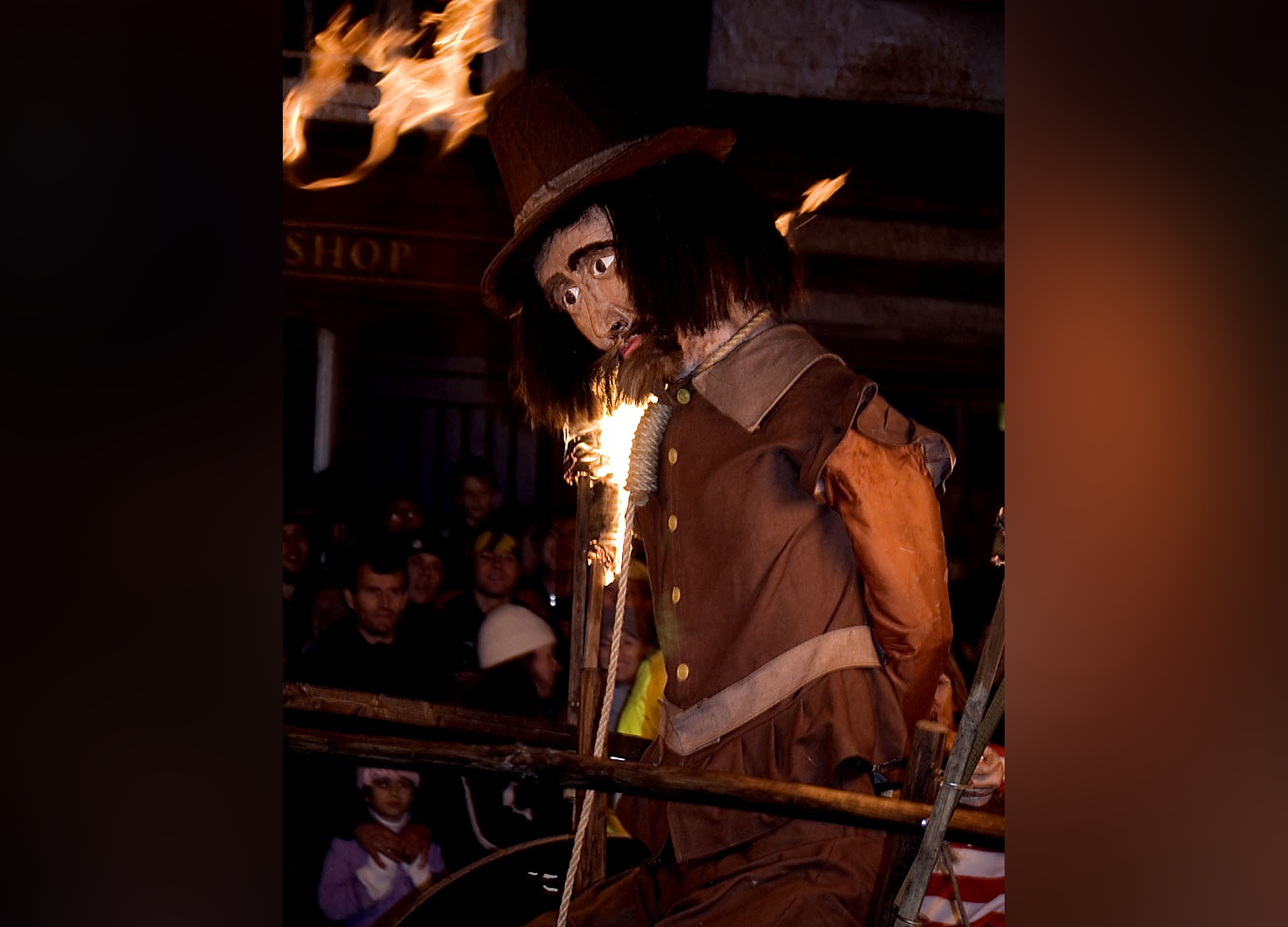 Zdjęcie przedstawia wielką kukłę mężczyzny. Kukła ubrana jest brązową kurtkę skórzaną, na głowie ma kapelusz, a na szyi założony powróz. Kukła ma głowę lekko pochyloną, brodę i wąsy, smutny wyraz twarzy, ciemne włosy do ramion. Przymocowana jest do drewnianych żerdzi. W tle widoczne postacie oraz płonące pochodnie.