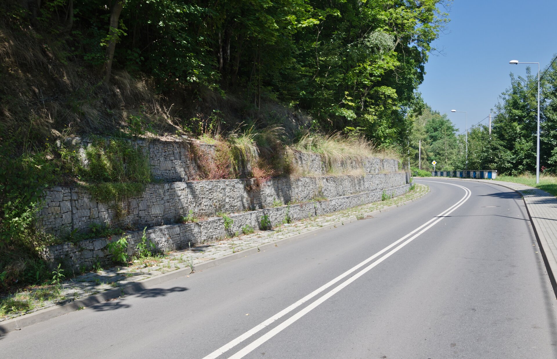 Zdjęcie przedstawia zbocze przy asfaltowej drodze.U podnóża zbocza znajduje się mur z kamieni. Mur ma trzy stopnie i zabezpiecza ziemię zbocza przed osunięciem się na drogę. 