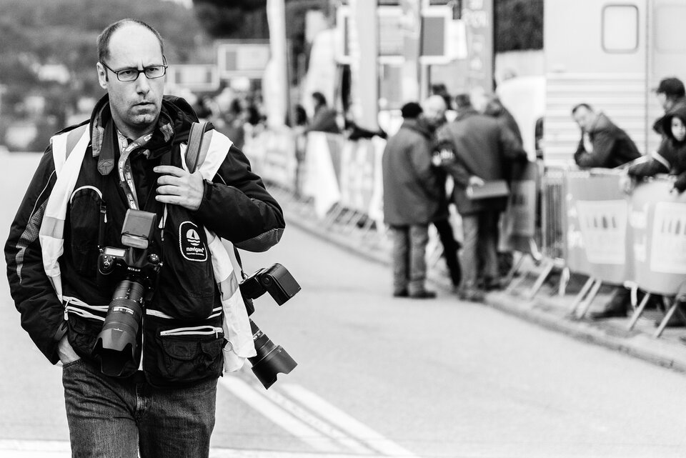 Na zdjęciu fotoreporter. Mężczyzna w średnim wieku w okularach idący ulicą. Ma dwa aparaty fotograficzne: jeden zawieszony na szyi, drugi na lewym ramieniu. Na plecach plecak. Po prawej stronie ulicy barierki, przy nich kilka osób.     