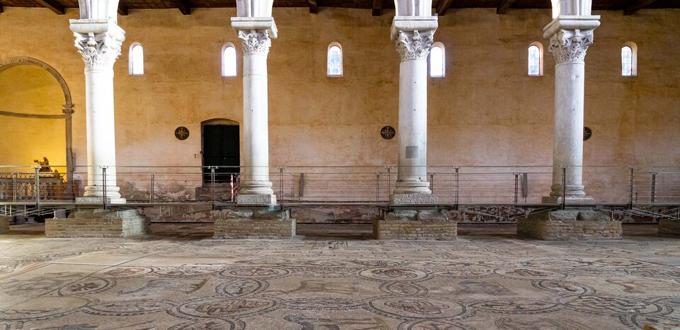 Fotografia przedstawia wnętrze budynku, którego konstrukcja została oparta na kolumnach z głowicami teodozjańskimi, które przypominają połączenie głowicy jońskiej z koryncką ale z większą ilością zdobień w postaci liści akantu. Kolumny stoją na ceglanych cokołach, które z kolei stoją na mozaice. Ściany budynku mają kolor beżowy, z lewej strony nisza, w której stoi figura kobiety trzymająca w dłoniach mężczyznę.