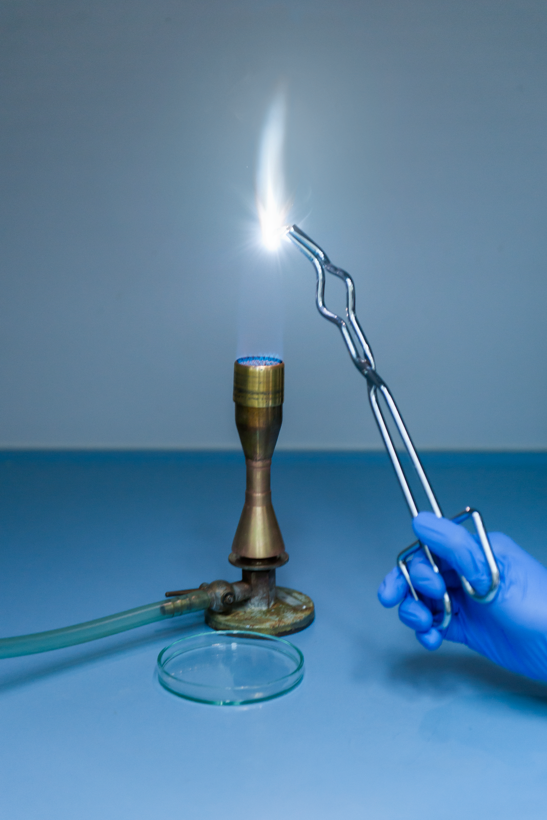 Zdjęcie przedstawia palnik gazowy, nad którym ręka eksperymentatora trzyma długie szczypce, których koniec znajduje się w zasięgu płomienia. Trzymana w szczypcach wstążka magnezowa spala się białym oślepiającym płomieniem.