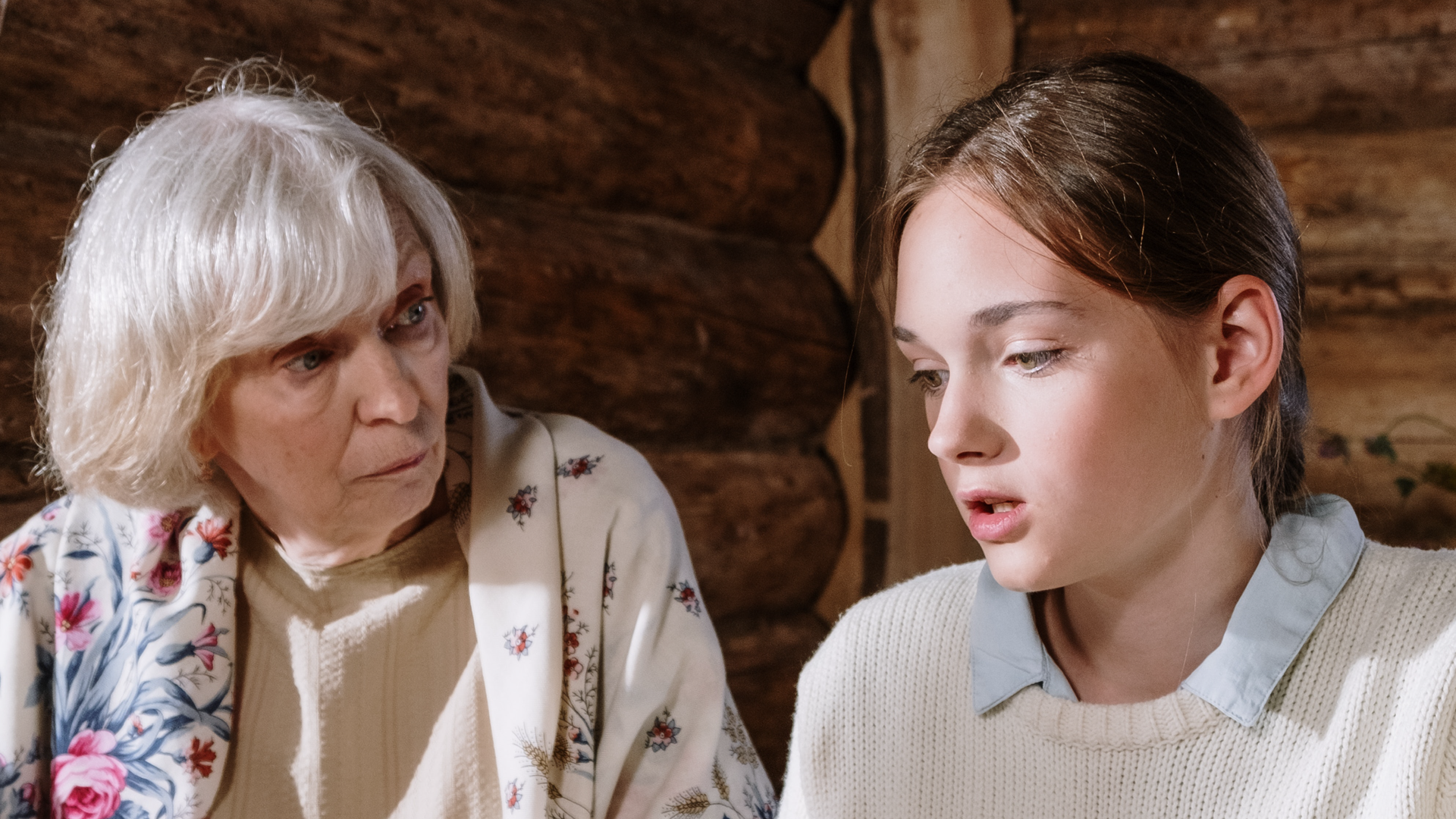 Zdjęcie przedstawia babcię i wnuczkę. Po lewej stronie znajduje się starsza, siwa kobieta. Kobieta nosi sweter w serek i wzorzystą kurtkę. Po prawej znajduje się młoda dziewczyna z włosami spiętymi z tyłu. Dziewczyna nosi bluzkę z kołnierzykiem i sweter.