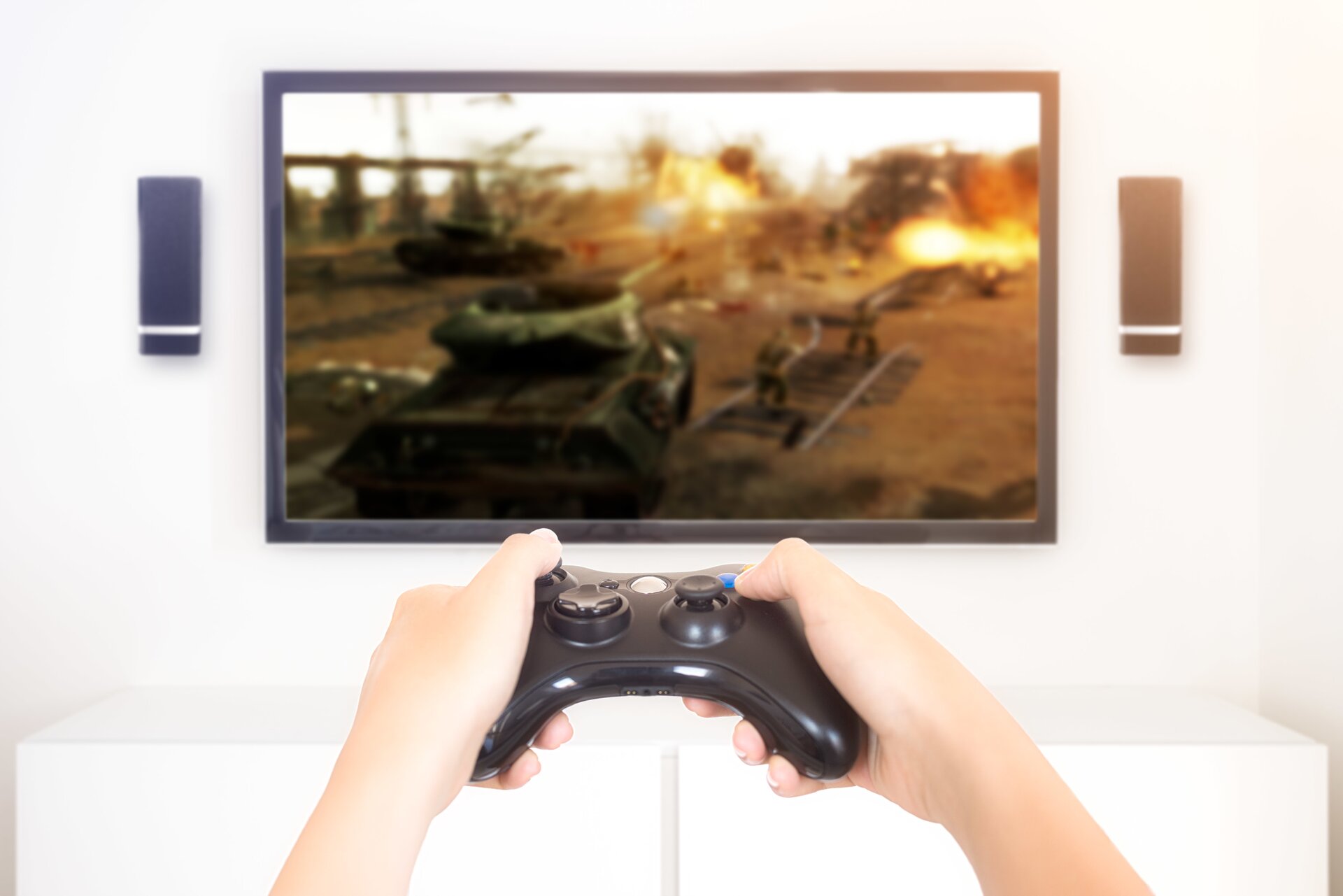 Fotografia przedstawia granie na konsoli. Widać tylko dłonie osoby trzymającej konsolę i duży ekran telewizora z grą o tematyce wojennej. Widoczny jest czołg, a dookoła niego znajdują się atakujący żołnierze. W tle rozbłyski i wybuchy.