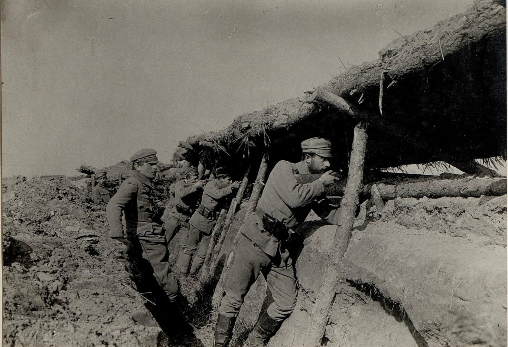 Zdjęcie przedstawia żołnierzy stojących w okopie. Nad nimi jest umocnienie z drewna i usypanej ziemi. Żołnierze celują z broni poprzez niewielką przestrzeń w nasypie.