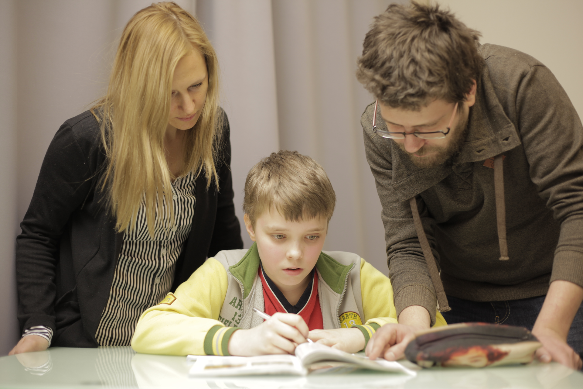 Fotografia prezentuje chłopca siedzącego przy stole i odrabiającego lekcje. Nad nim pochylają się rodzice pomagający mu odrabiać zadania.