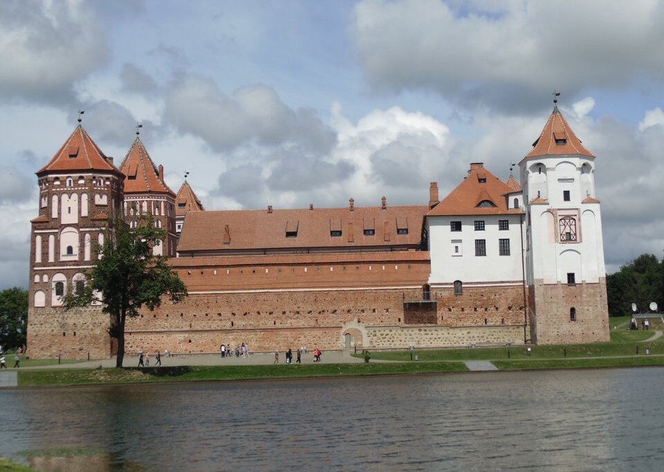 Fotografia przedstawiająca turystyczną atrakcję Białorusi - Zamek w Mirze. Zamek zbudowany jest z drobnej cegły , po bokach zamku widnieją 4 wierze. Zamek otoczony jest murem. Znajduje się nad rzeką widoczną na pierwszym planie ilustracji.