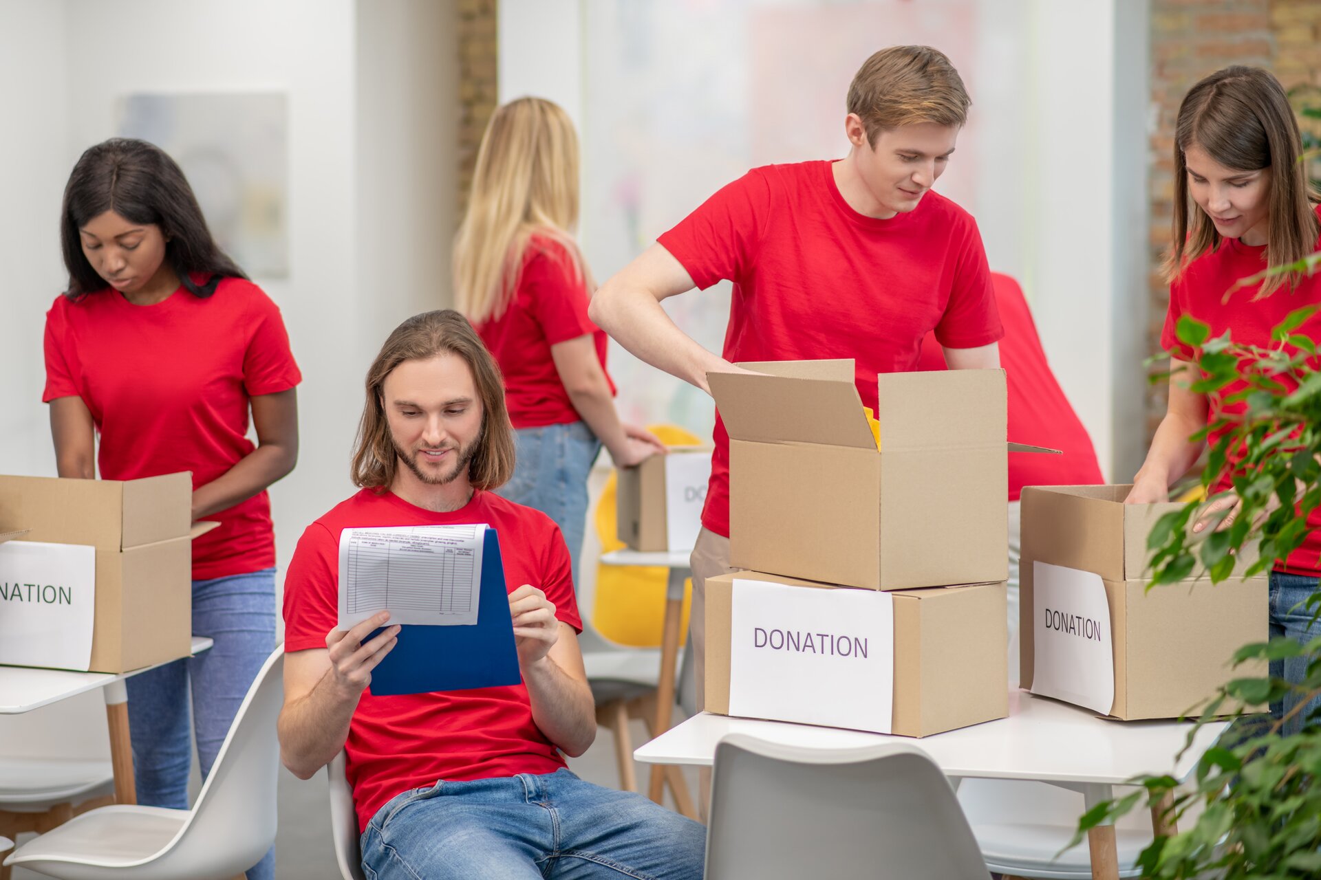 Zdjęcie przedstawia grupę młodych ludzi, ubranych w czerwone koszulki, stojących przy stolikach i wkładających coś do kartonów. Na kartonach widoczne są duże białe kartki z napis z czarnych liter w języku angielskim:DONATION. Z lewej strony widoczny jest młody mężczyzna, który siedzi i trzyma w rękach niebieską podkładkę z przypiętą kartką. Spogląda na nią i prawą ręką dotyka podkładki, wykonując gest sprawdzenia. 