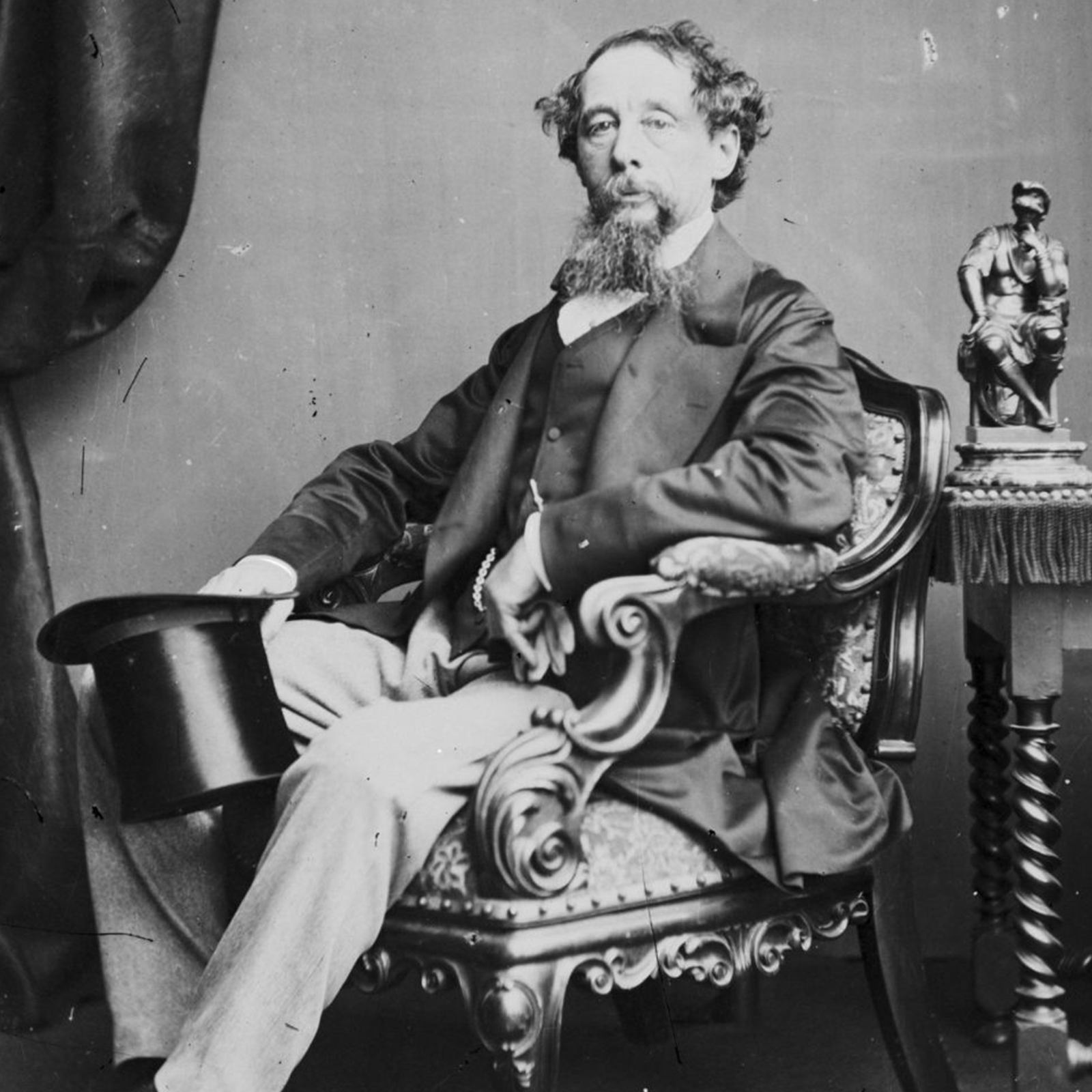 Czarno‑białe zdjęcie przedstawia siedzącego w fotelu mężczyznę w średnim wieku, zwróconego lekko w lewo, z twarzą w kierunku obiektywu. Mężczyzna ma krótkie, kręcone, ciemne włosy, dłuższą brodę oraz wąsy. Jest ubrany w ciemną marynarkę i kamizelkę, spod której wystaje biała koszula, oraz jasne spodnie. W prawej dłoni, opartej na poręczy fotela, trzyma czarny cylinder. Lewą rękę ma opartą o poręcz i przyciągniętą do brzucha, a w dłoni trzyma rękawiczkę. Fotel ma zdobione, drewniane poręcze i jest obity materiałem. Na prawo od fotela widoczna jest mała rzeźba stojąca na stoliczku, a po lewej stronnie fragment ciemnej, upiętej zasłony.